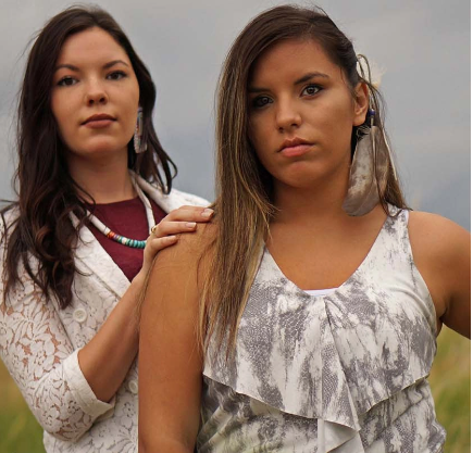 Two women stand outdoors; one in a white patterned top with a feather earring, the other in a lace jacket with turquoise jewelry, resting a hand on the other's shoulder. Two women stand outdoors; one in a white patterned top with a feather earring, the other in a lace jacket with turquoise jewelry, resting a hand on the other's shoulder.