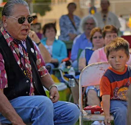 An older man speaks while sitting outdoors next to a young boy on a chair, with a group of people seated in the background.